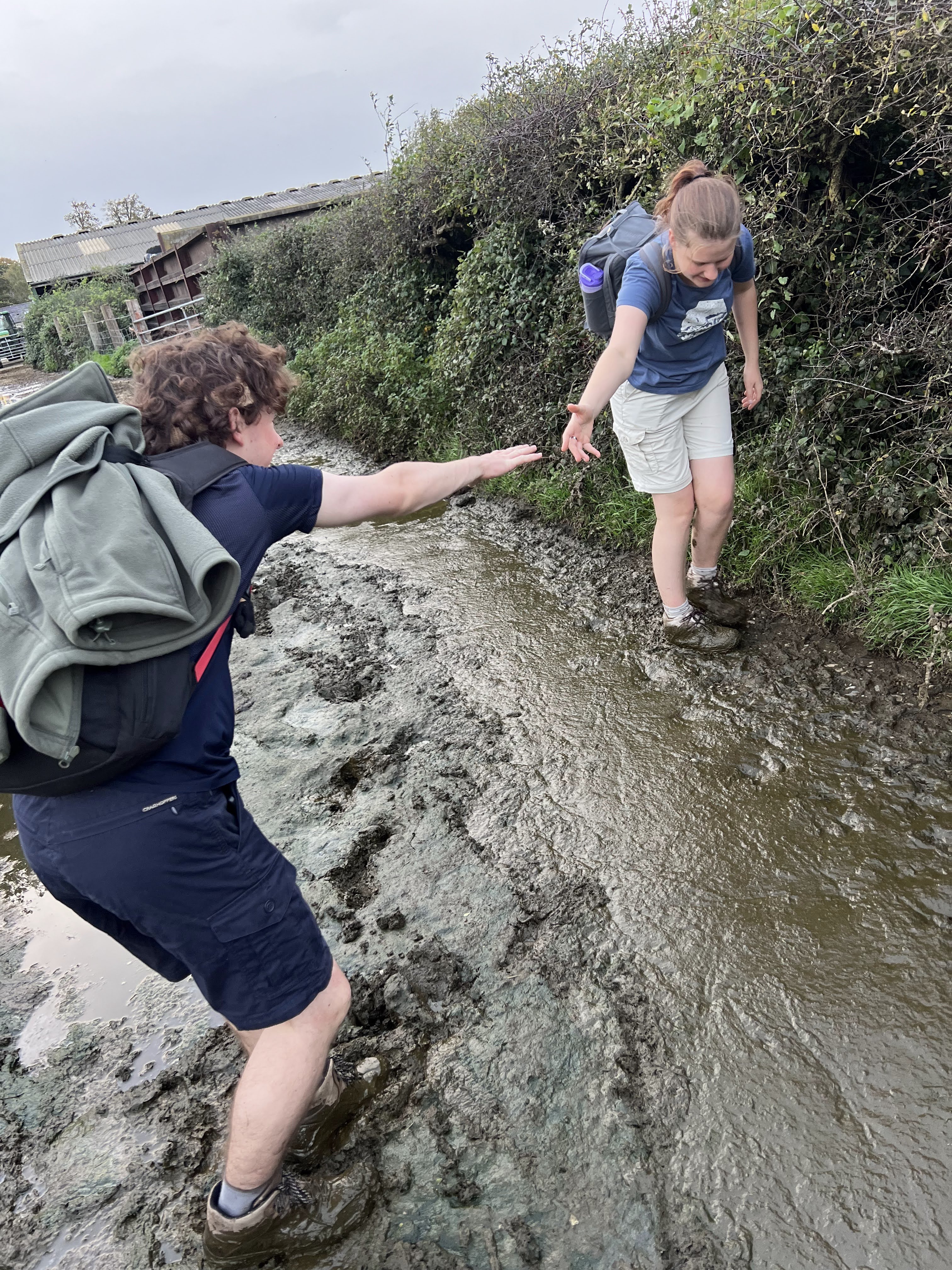 Nick in the cow poop reaching over to Mya to give her a hand as she is precariously balanced on the edge